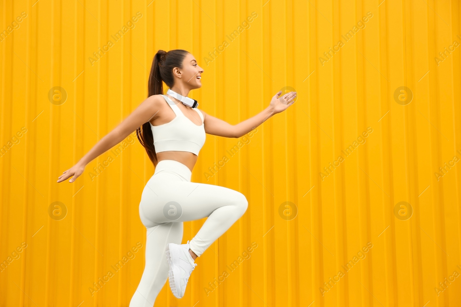 Photo of Young woman in sportswear with headphones  running near corrugated yellow metal wall. Space for text