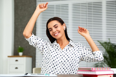 Photo of Happy young woman listening to music through wireless earphones at table in office