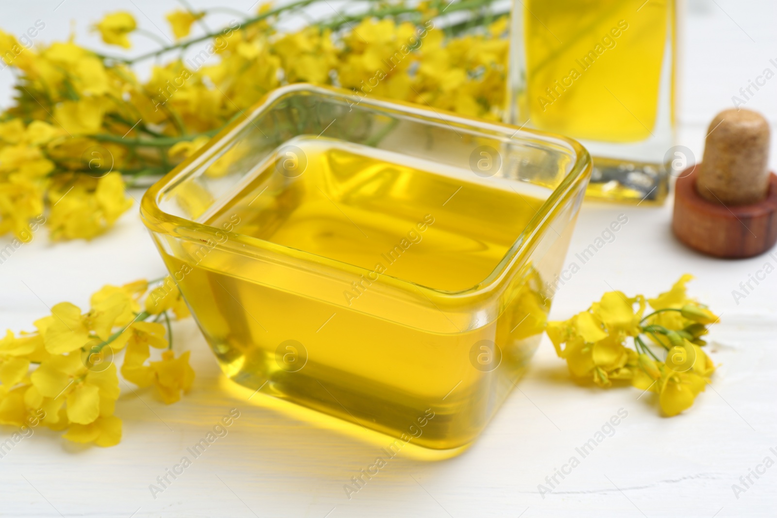 Photo of Rapeseed oil in glass bowl and beautiful yellow flowers on white wooden table, closeup
