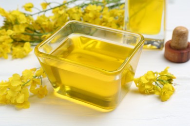 Rapeseed oil in glass bowl and beautiful yellow flowers on white wooden table, closeup