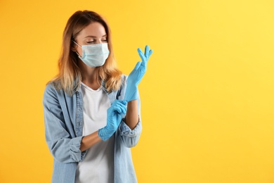 Young woman in protective mask putting on medical gloves against yellow background. Space for text