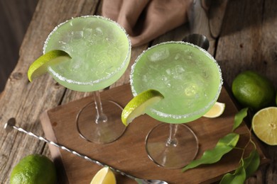 Photo of Delicious Margarita cocktail in glasses, limes, green leaves and bar spoon on wooden table