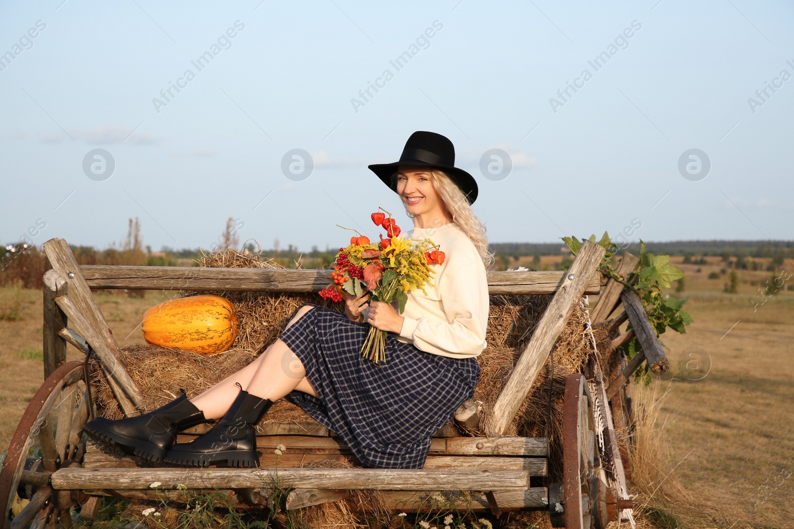 Photo of Beautiful woman with bouquet sitting on wooden cart with pumpkin and hay in field. Autumn season