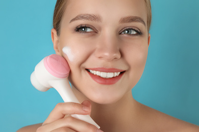 Young woman washing face with brush and cleansing foam on light blue background. Cosmetic products