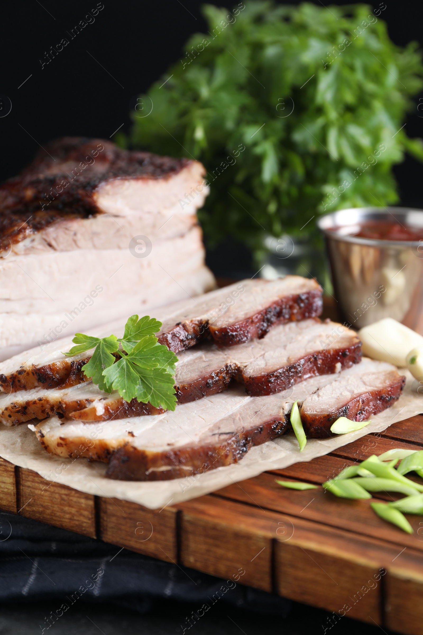 Photo of Pieces of baked pork belly served with sauce and parsley on black textured table, closeup