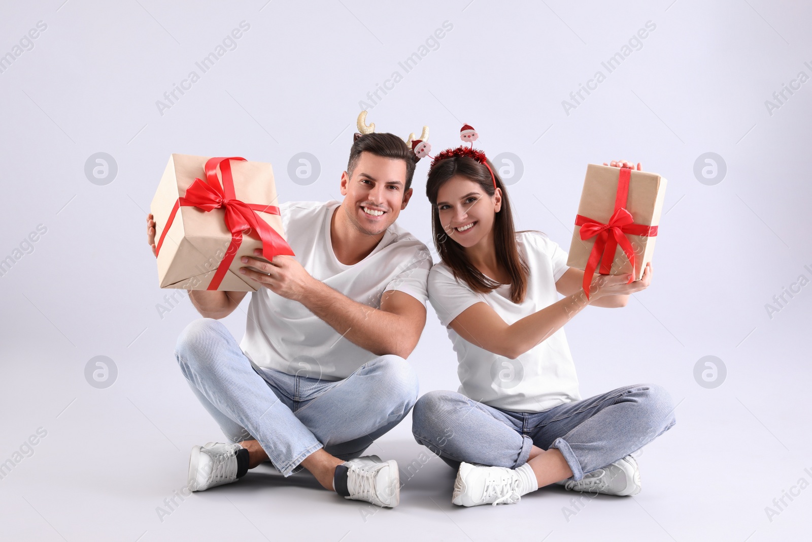 Photo of Beautiful happy couple in Christmas headbands  sitting with gifts on light background