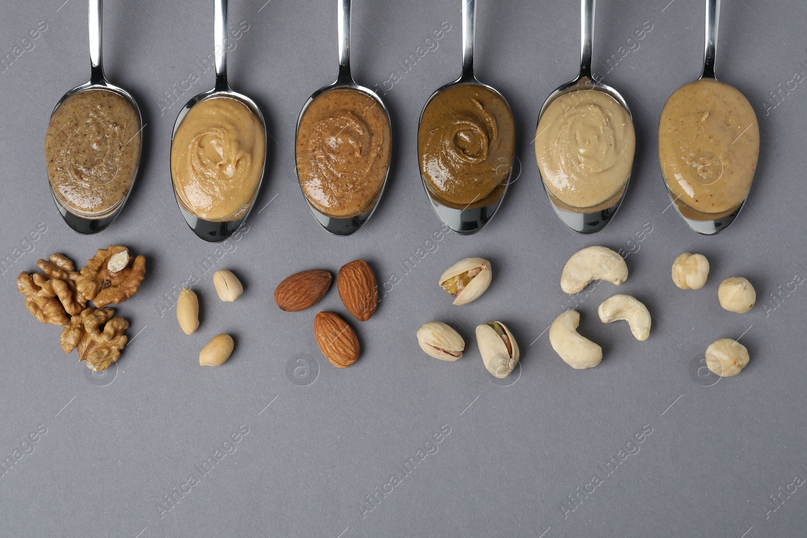 Photo of Tasty nut butters in spoons and raw nuts on gray table, flat lay. Space for text