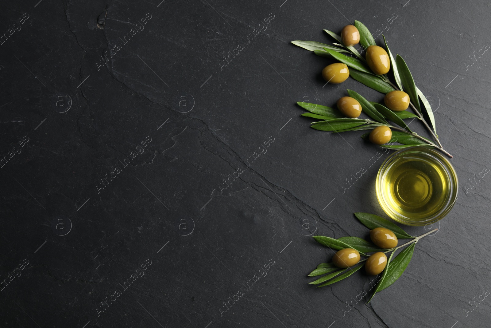 Photo of Cooking oil in bowl, olives and leaves on black textured table, flat lay. Space for text