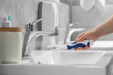 Photo of Woman washing electric toothbrush under flowing water from faucet in bathroom, closeup