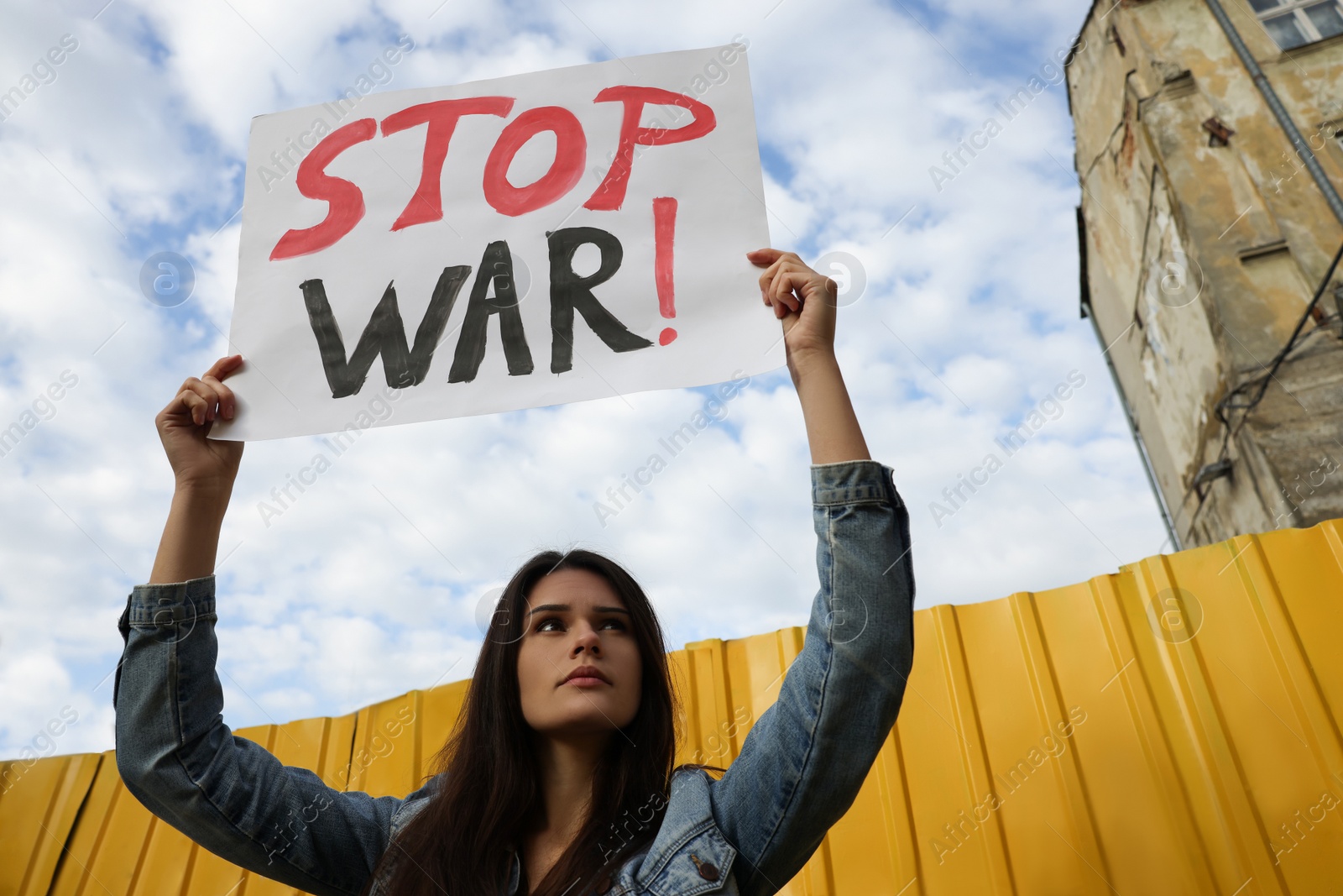 Photo of Sad woman holding poster Stop War near yellow fence under blue cloudy sky