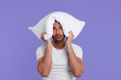 Photo of Tired man covering ears with pillow on violet background. Insomnia problem