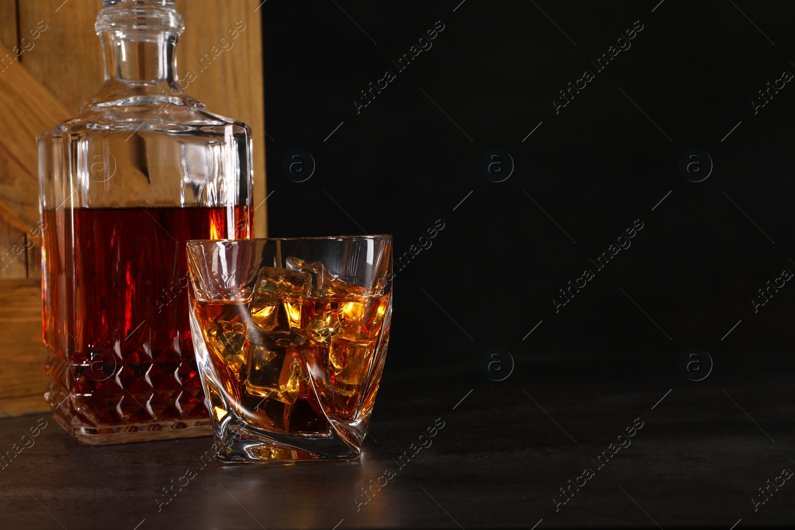 Photo of Whiskey in glass and bottle near wooden crate on dark table against black background. Space for text