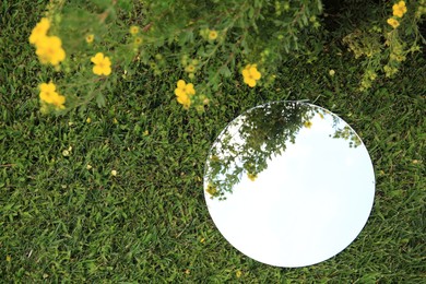 Photo of Round mirror on green grass reflecting sky and beautiful flowers, top view