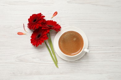 Photo of Cup of coffee and red gerbera flowers on white wooden table, flat lay