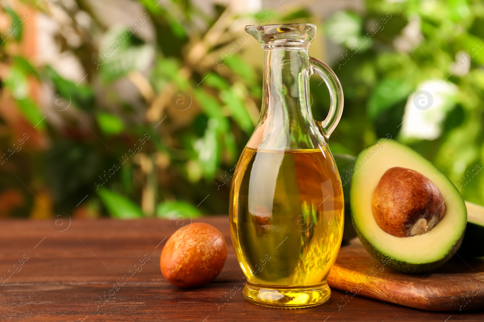 Photo of Glass jug of cooking oil and fresh avocados on wooden table against blurred green background, space for text