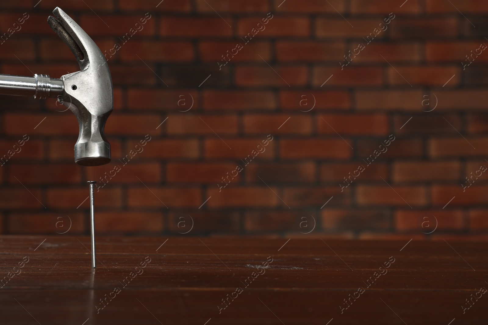 Photo of Hammering metal nail on wooden table against brick wall. Space for text