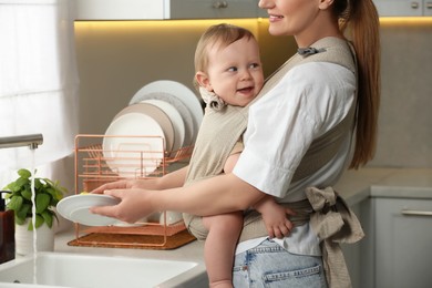 Photo of Mother holding her child in sling (baby carrier) while washing plates in kitchen