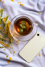 Photo of Cup of aromatic herb tea, smartphone and dry autumn leaves on white cloth, flat lay