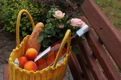 Yellow wicker bag with roses, peaches, baguette and wine on wooden bench outside. Space for text