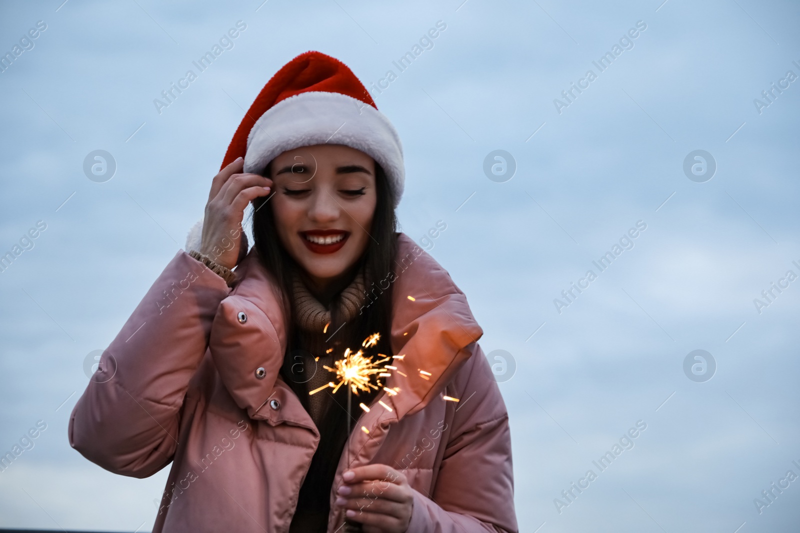 Photo of Woman in Santa hat and warm clothes holding burning sparkler outdoors