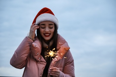 Photo of Woman in Santa hat and warm clothes holding burning sparkler outdoors