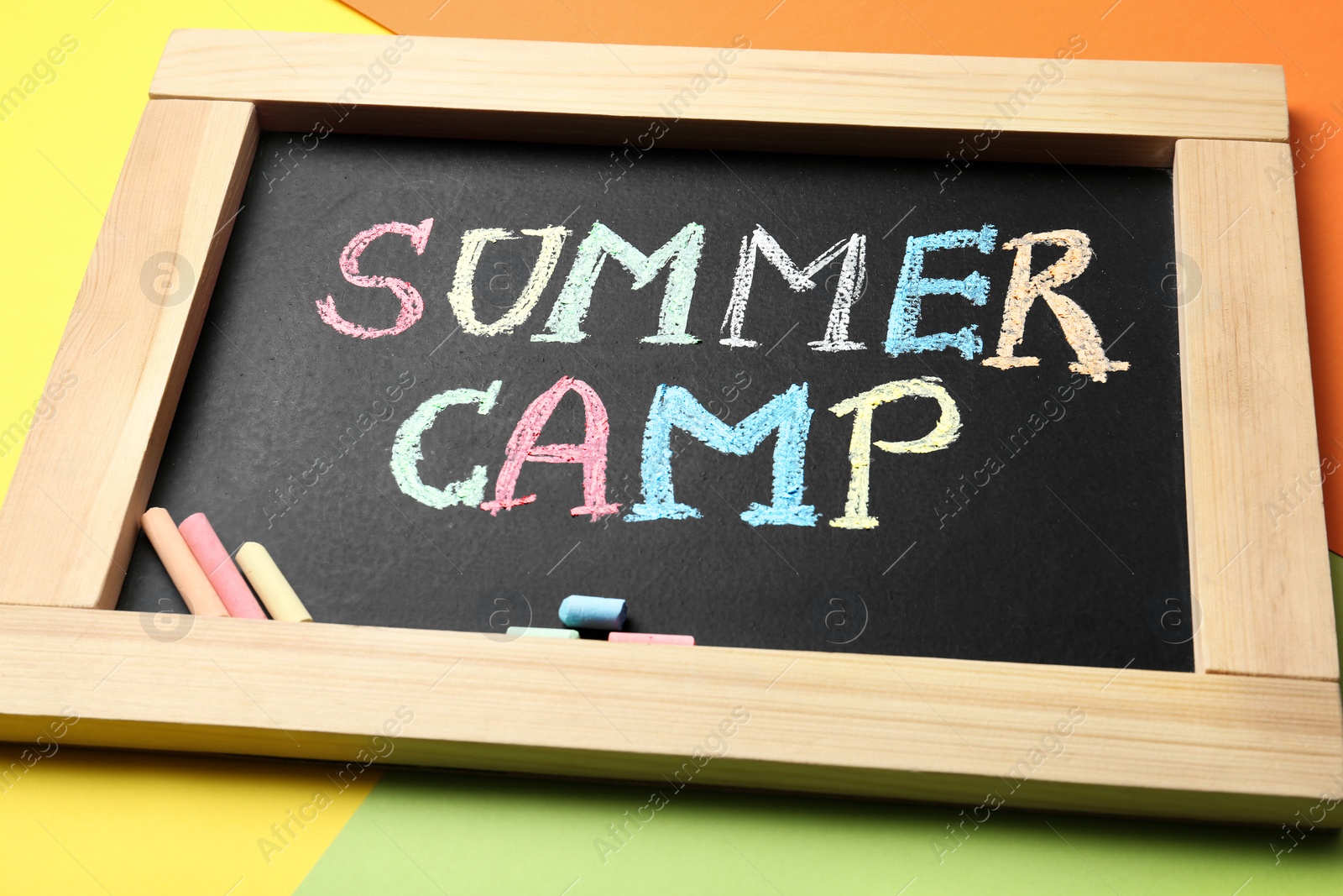 Photo of Small blackboard with text SUMMER CAMP and chalk sticks on color background