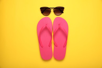 Photo of Stylish pink flip flops and sunglasses on yellow background, flat lay