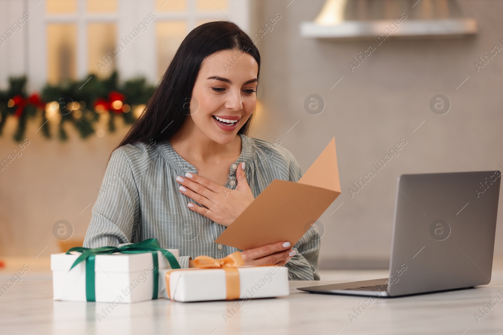 Photo of Celebrating Christmas online with exchanged by mail presents. Smiling woman reading greeting card and gifts during video call at home