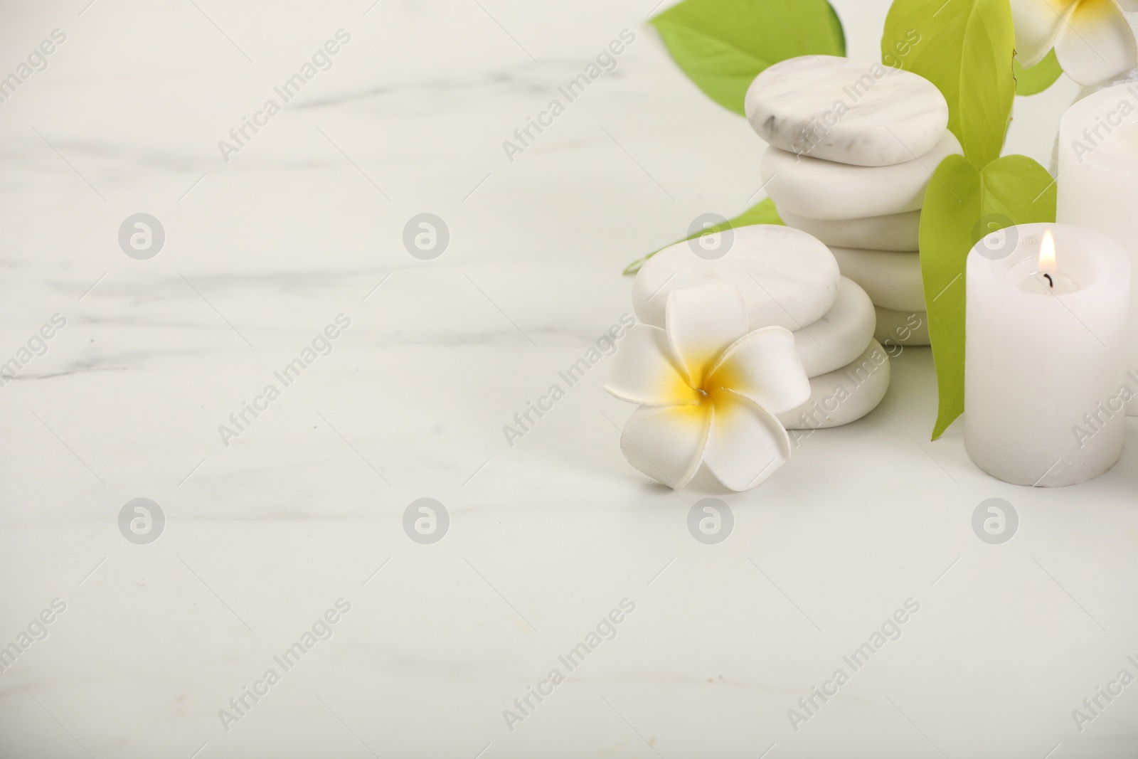 Photo of Spa composition. Burning candle, plumeria flower, stones and green leaves on white marble table, space for text