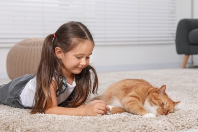 Photo of Smiling little girl petting cute ginger cat on carpet at home