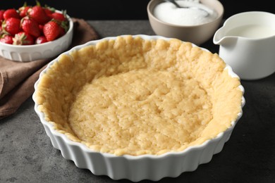 Photo of Making shortcrust pastry. Raw dough in baking dish, strawberries, sugar and milk on grey table