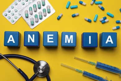Word Anemia made with blue wooden cubes, pills, syringes and stethoscope on yellow background, flat lay
