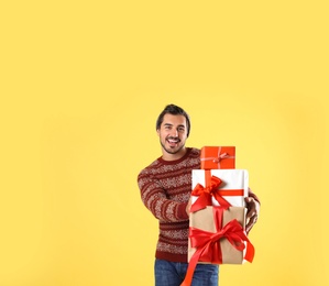 Happy man in Christmas sweater and hat holding gift boxes on yellow background