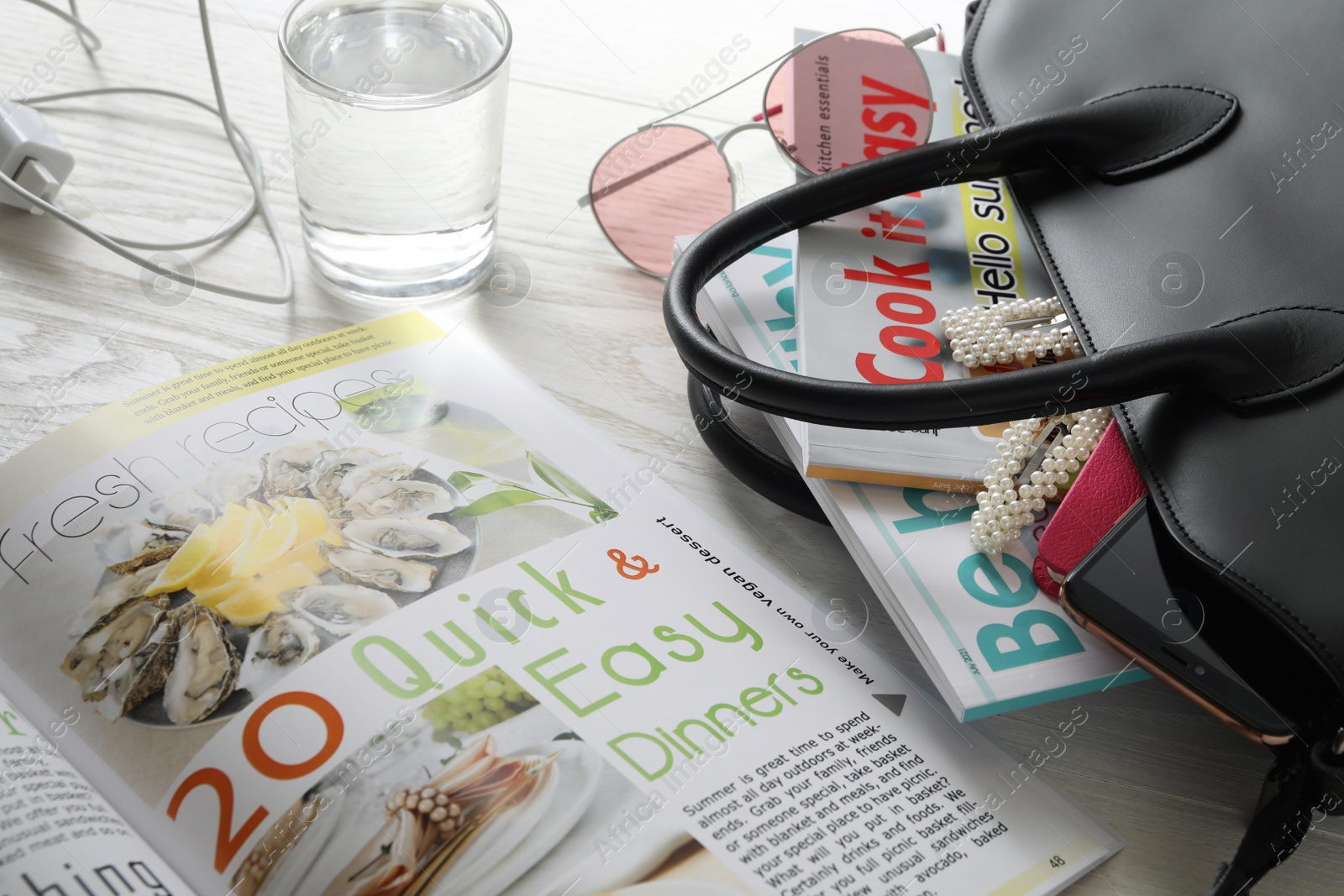 Photo of Bag with different magazines, accessories and glass of water on white wooden table, closeup