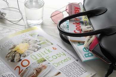 Bag with different magazines, accessories and glass of water on white wooden table, closeup