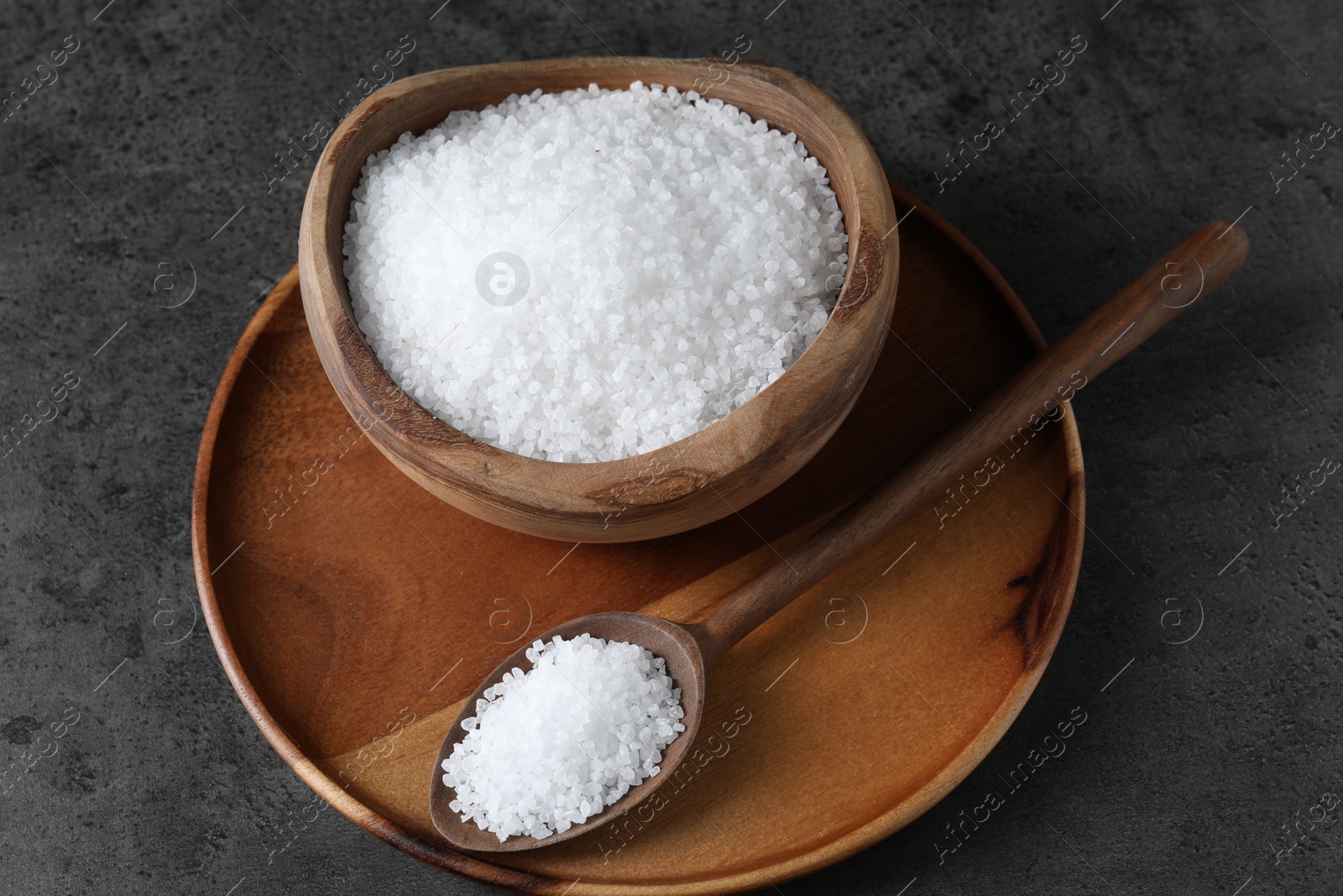 Photo of Natural salt in wooden bowl and spoon on dark grey table