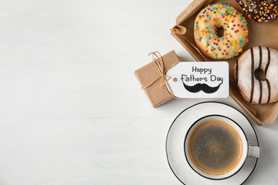 Photo of Tag with phrase HAPPY FATHER'S DAY, donuts, coffee and gift on white wooden table, flat lay. Space for text