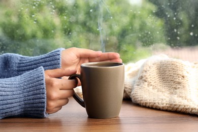 Woman with cup of hot drink at wooden table near window on rainy day, closeup