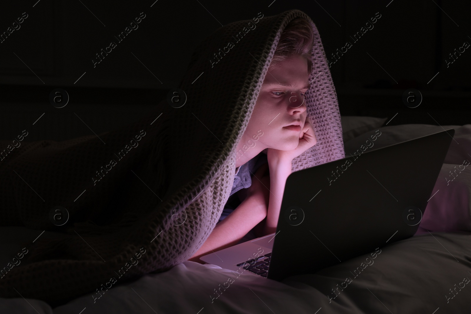 Photo of Teenage boy using laptop under blanket on bed at night. Internet addiction