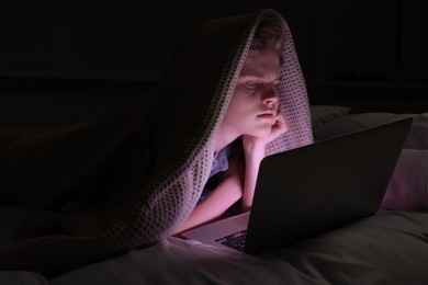 Photo of Teenage boy using laptop under blanket on bed at night. Internet addiction