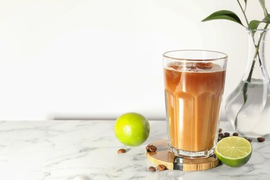 Photo of Refreshing iced coffee with milk in glass, beans and fresh limes on white marble table, space for text