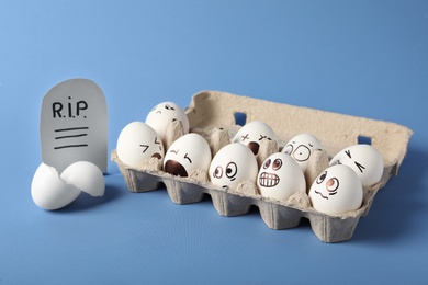 Photo of Eggs with drawn faces and paper grave near broken shell on blue background. April fool's day