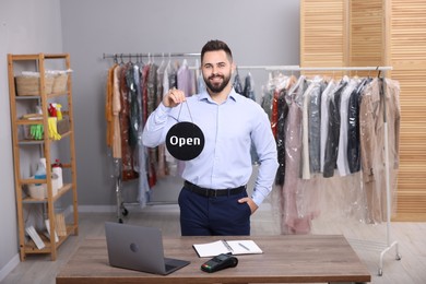 Dry-cleaning service. Happy worker holding Open sign at workplace indoors