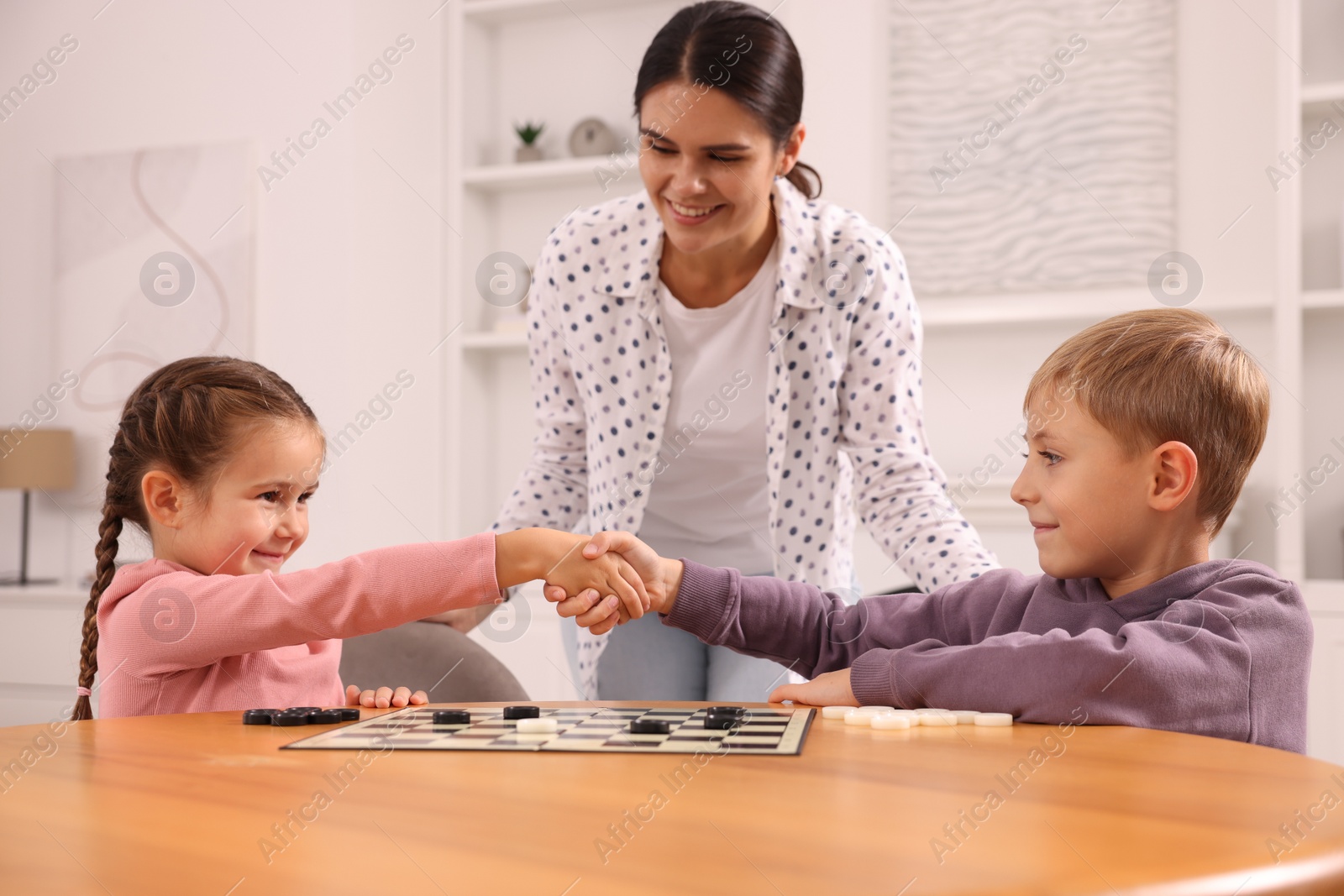 Photo of Family playing checkers at wooden table in room. Children shaking hands after game