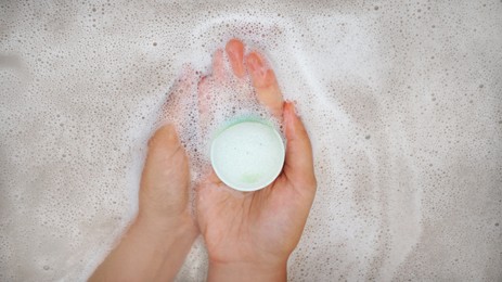 Photo of Woman holding bath bomb over water with foam, top view. Space for text