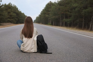 Young woman with backpack sitting on road near forest, back view