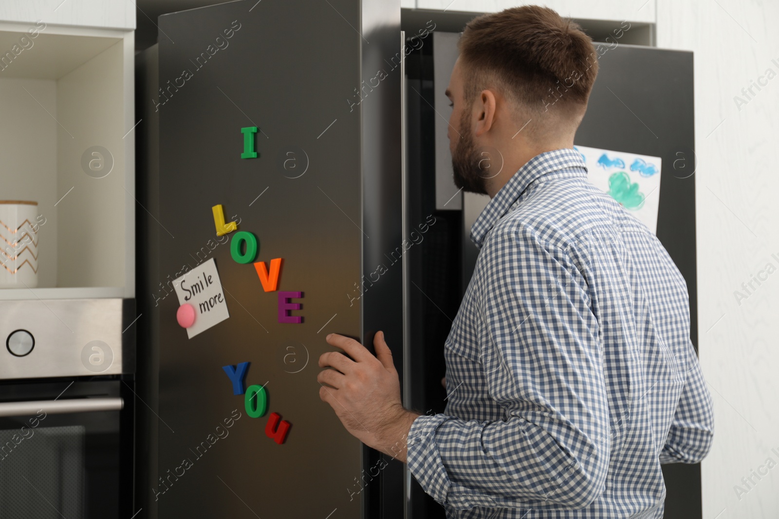 Photo of Man opening refrigerator door with notes and magnets in kitchen