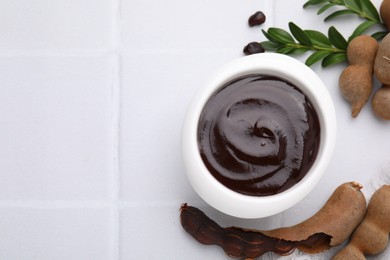 Photo of Bowl of tasty tamarind sauce, leaves and fresh pods on white tiled table, flat lay. Space for text
