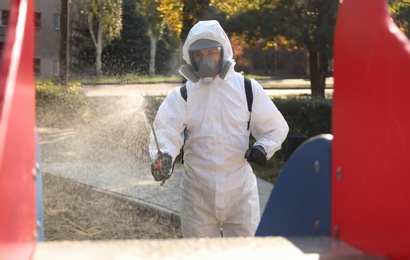 Photo of Man in hazmat suit spraying disinfectant onto slide at children's playground. Surface treatment during coronavirus pandemic