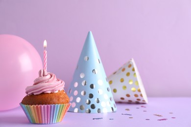Photo of Delicious birthday cupcake with candle near party hats  and balloon on violet background, space for text